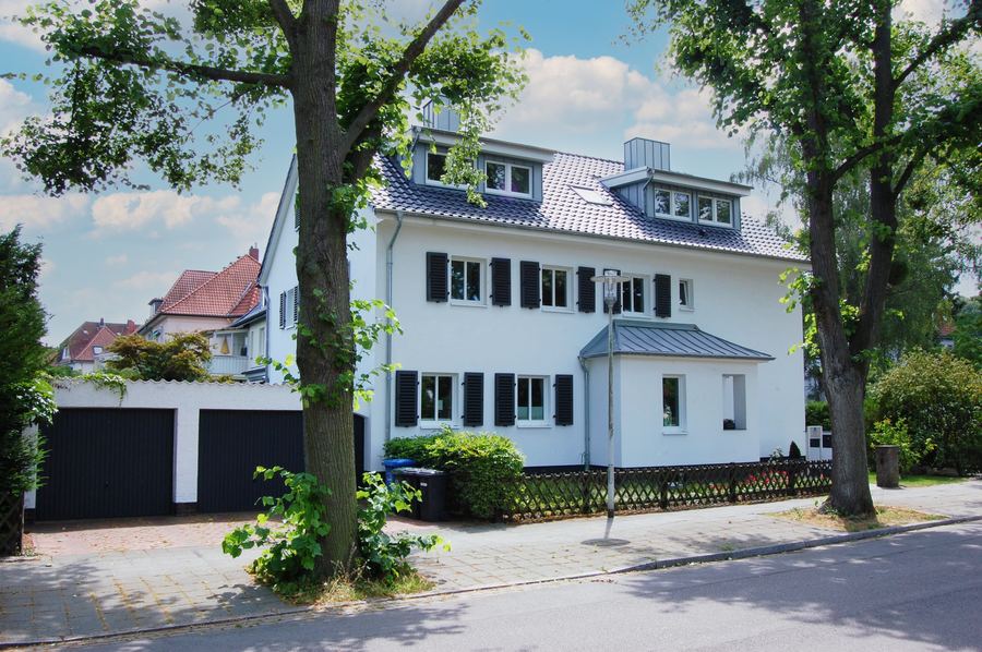 Weißes Wohnhaus mit dunklen Klappläden hinter alten Bäumen an der Straße