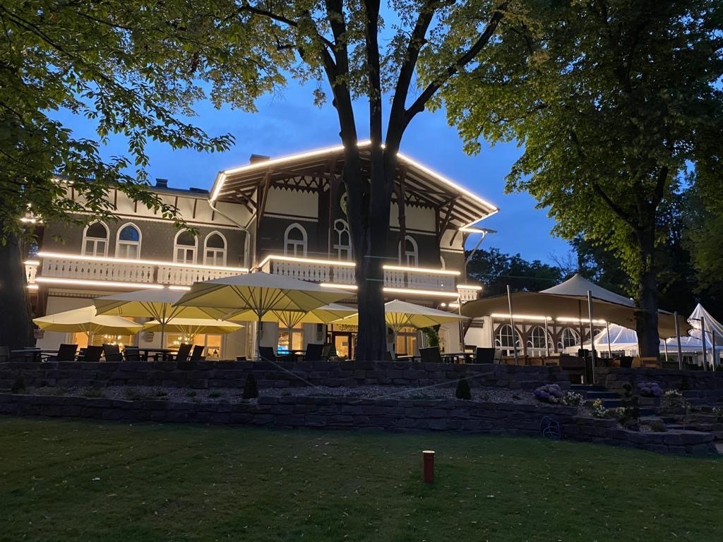 Saniertes Denkmalgebäude in der Abenddämmerung: beleuchtete Holzveranda, Spitzbogenfenster und Terrasse mit Sonnenschirmen