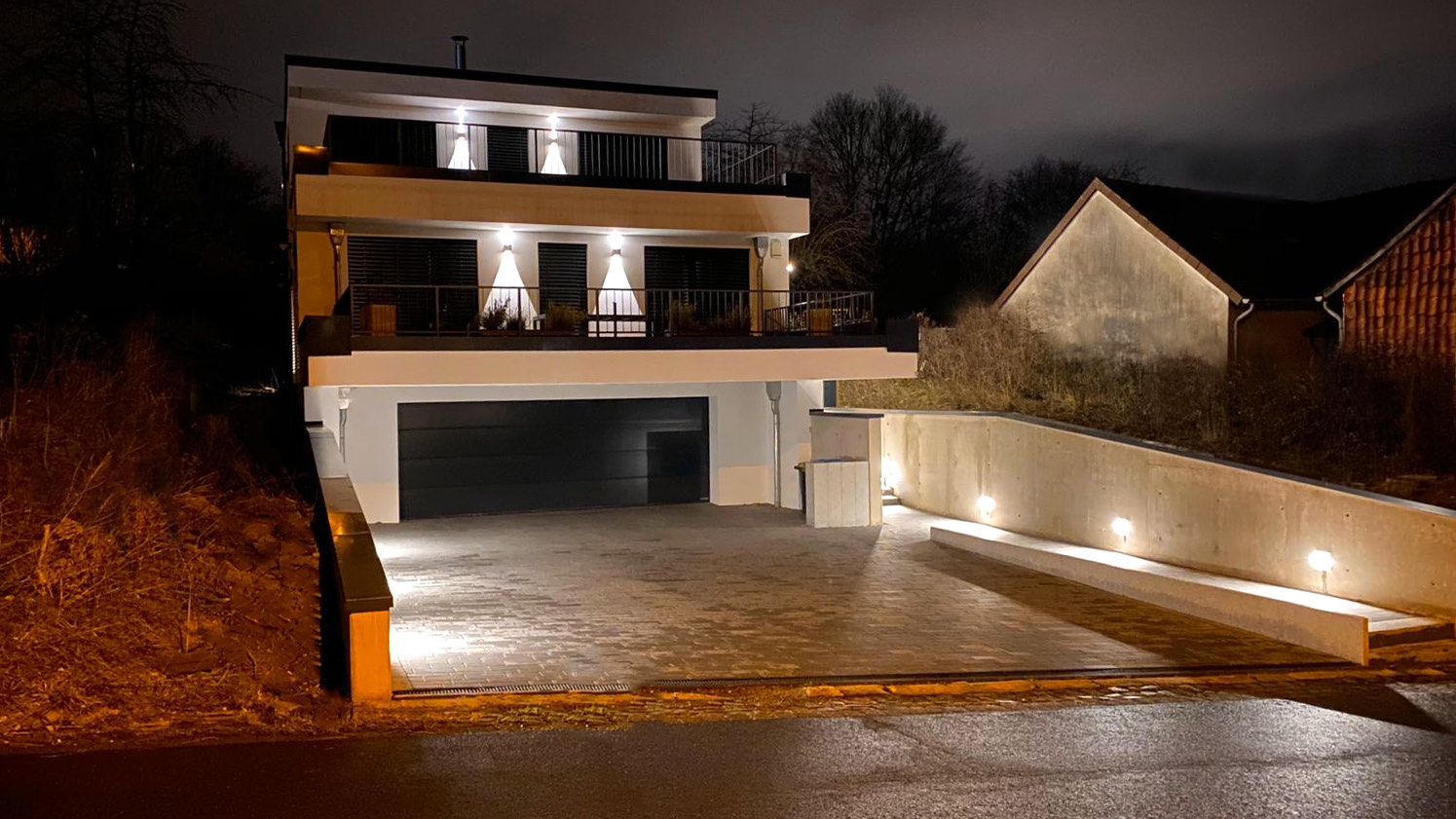 Das fertiggestellte Haus am Hang bei Nacht: beleuchtete Terrassen über der Garage, Stützmauer und gepflasterte Zufahrt