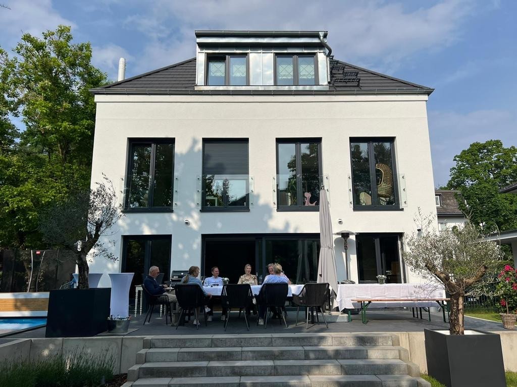 Gartenseite eines Einfamilienhauses: weiß verputzte Fassade mit bodentiefen Fenstern, Terrasse mit langer Tafel und Pool im Vordergrund
