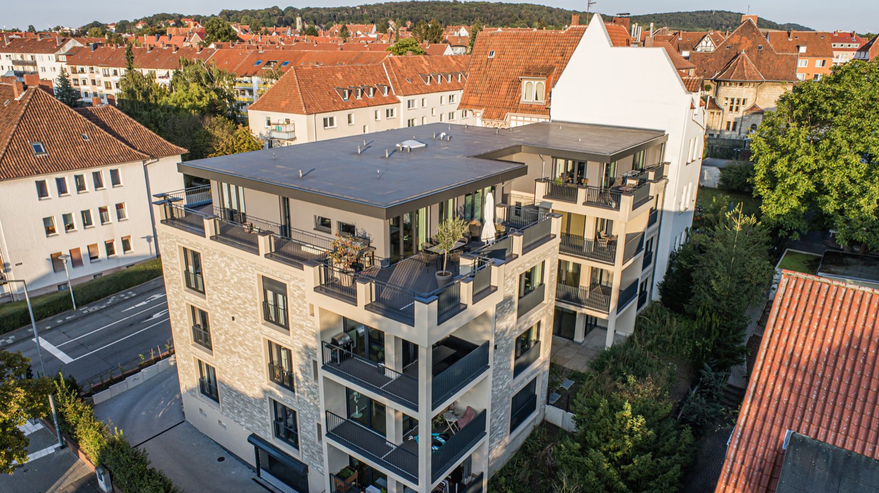 Luftaufnahme eines fertiggestellten Mehrfamilienhauses mit heller Natursteinfassade, umlaufenden Balkonen und begrünter Dachterrasse